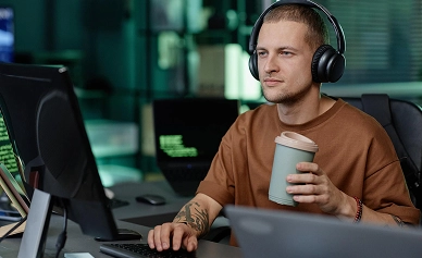 Een geconcentreerde man met een koptelefoon op, zittend achter een computer met een kop koffie, terwijl hij verschillende taken tegelijk uitvoert in een moderne werkruimte met digitale schermen